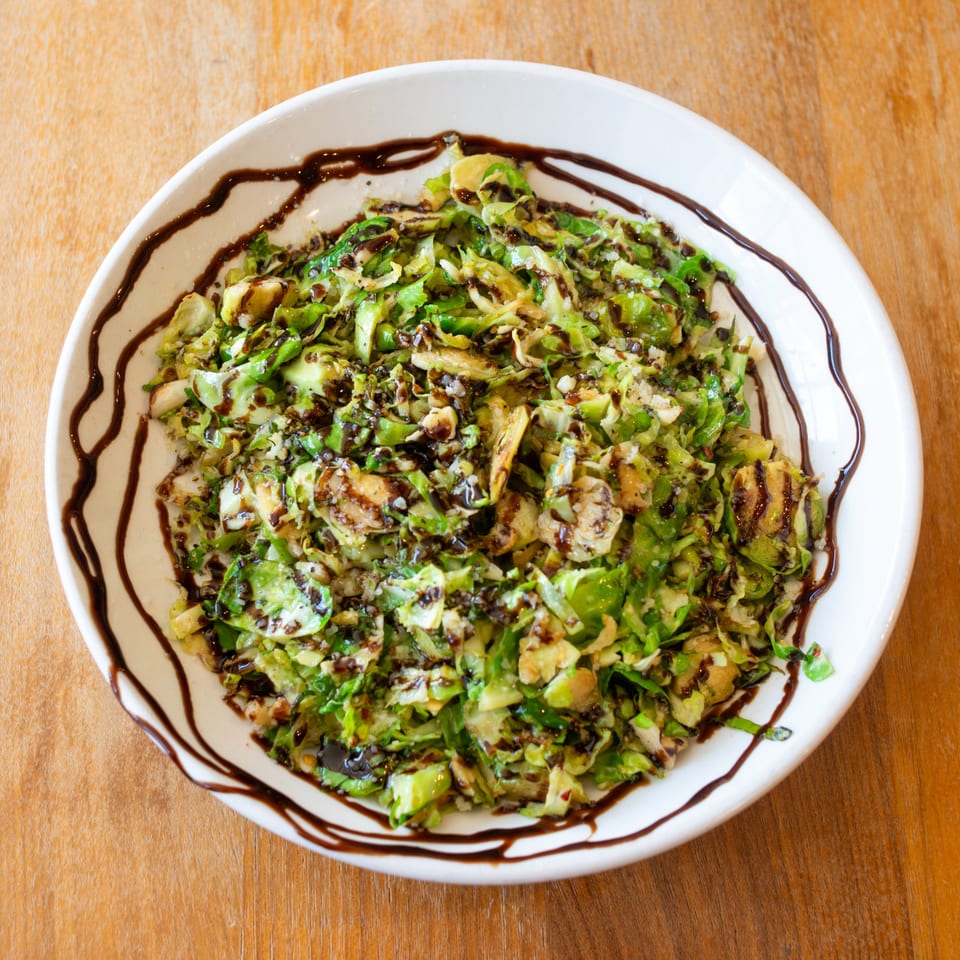 A white bowl of shredded Brussels sprouts with balsamic glaze and parmesan cheese on a wooden table.