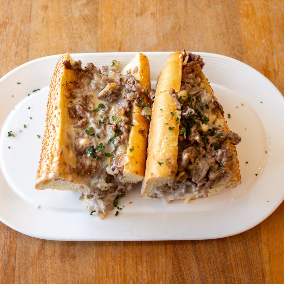 Two halves of a cheesy steak and mushroom sub sandwich on a white plate.