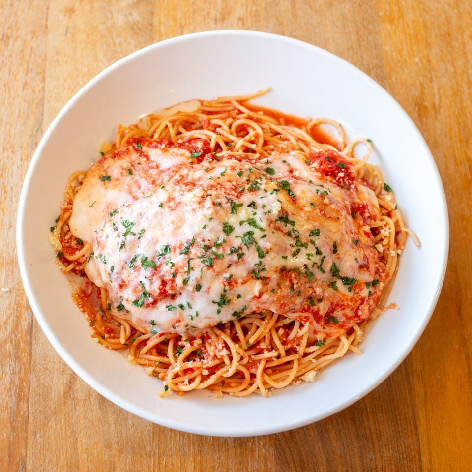 Chicken Parmesan with spaghetti, red sauce, melted mozzarella, and parsley in a white bowl.