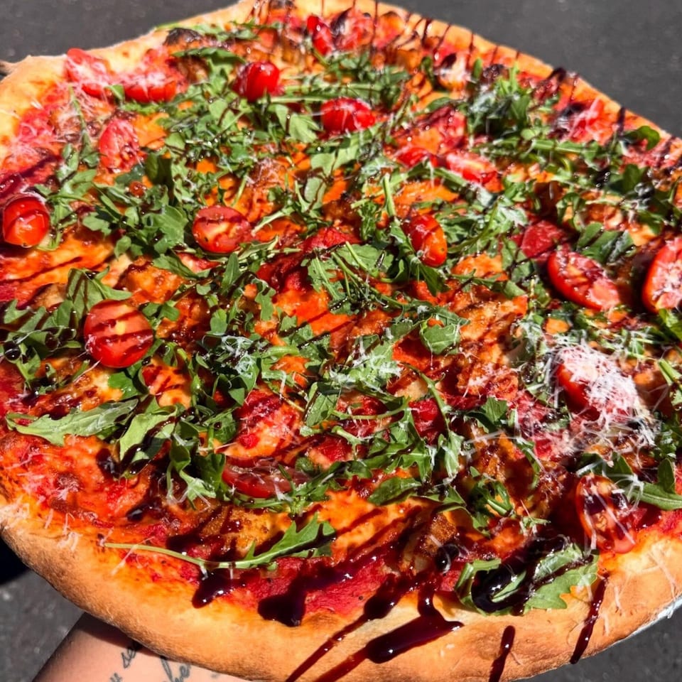 A Pizza Wheel Brick Oven pizza with arugula, cherry tomatoes, cheese, and balsamic glaze.