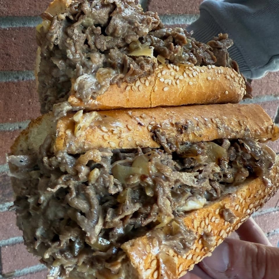 Two stacked, overflowing cheesesteak sandwiches with melted cheese and onions on sesame seed rolls.