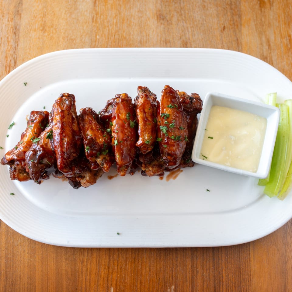 A white platter of BBQ chicken wings with a side of ranch dressing and celery sticks.